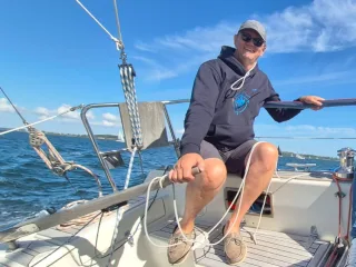 Ein Mann mit Sonnenbrille und Hoodie sitzt im Cockpit eines Segelboots und steuert mit der Pinne.
